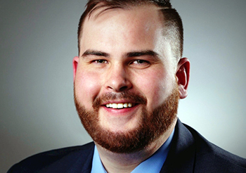 Photo of Brett. He has red hair and a beard, is wearing a dark blue coat, light blue shirt, and colorful paisley tie.