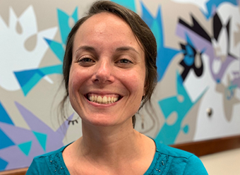 Photo of Colleen smiling. She has brown hair and is wearing a blue short. She stands in front of an abstract wall mural that is beige, blue, teal, black and white.