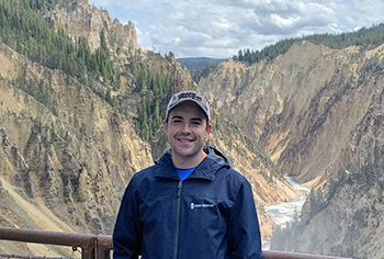 photo of Dan standing on a bridge over a mountain gorge with snow still in the shadows, water rushes below and sunny blue skies above