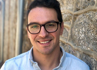 close up of Igor, smiling, he has black har parted ont he side and is wearing glasses and a blue shirt. He is leaning against a stone wall.
