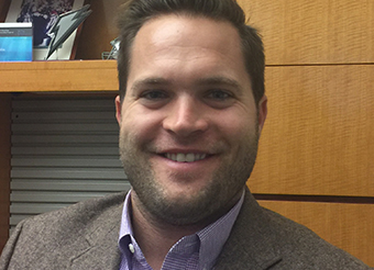 close up photo of John, smiling. He has blond hair and is wearing a purple shirt and a gray sportscoat.