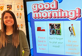 photo of Kim, standing with a big smile, in front of a classroom smartboard that welcomes the students