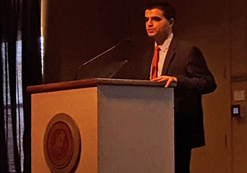 photo of Neil in a dark suit and red tie, standing at a posium on stage.