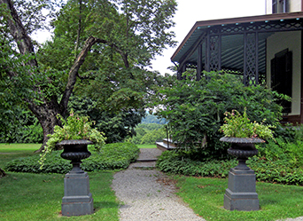 photo of the south porch at Locust Grove. It shows wrought iron pillars, and a high celing that frame views of the Hudson River in the distance. There are two urns with potted flowers which flank a gravel walk that approaches the porch stairs.