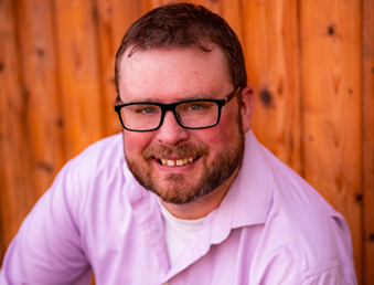Photo of Ross, seating and smiling. He has red hair and a short beard, wears black-rimmed glasses and a purple button-down shirt.