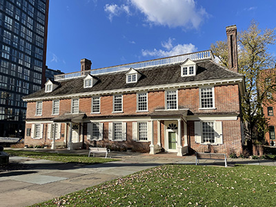 exterior photo of a red brick building, 2 stories + mansard roof with dormers. There is a lawn with benches surrounding the building and a high-rise next door.