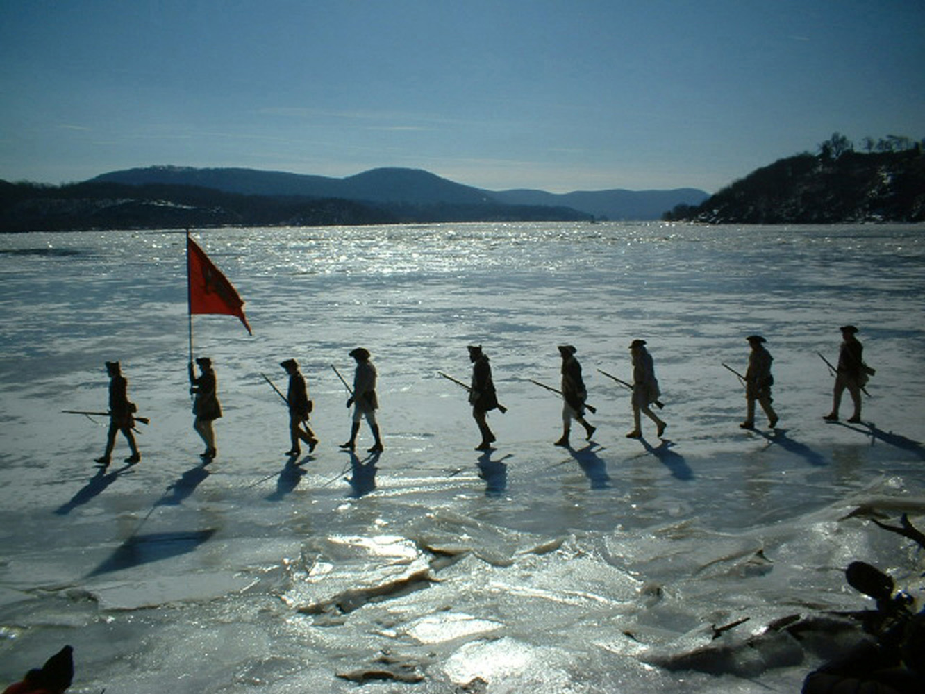 Nine men in Revolutionary era costumes, with a flag and rifles, walking across the frozen Hudson River