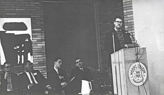 black and white photo of a man with glasses speaking at a pudium with four men in suits seated behind