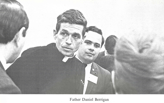 black and white photo of someone speaking to Father Daniel Berrigan in a crowded room 