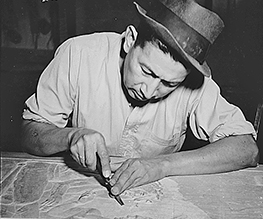 Kidd Smith, Seneca carver, working on a traditional carving. He wears a white button-down shirt and a fedora.