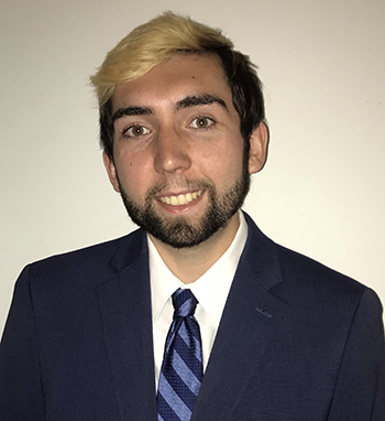 Close up photo of Aaron wearing a blue suit with a white shirt and a striped blue tie. His hair and beard is black, but his bangs are bleached blond. He is smiling. 