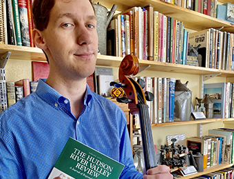 Photo of Alex standing in front of a library wall of books and artifacts. He is wearing a blue shirt and tan khakis. He holds his cello in one hand and a copy of the Spring 2022 issue of The Hudson River Valley Review in the other.