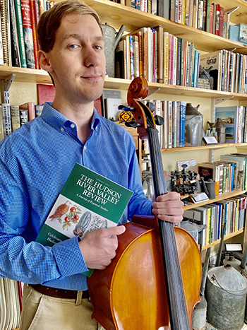 Photo of Alex standing in front of a library wall of books and artifacts. He is wearing a blue shirt and tan khakis. He holds his cello in one hand and a copy of the Spring 2022 issue of The Hudson River Valley Review in the other.