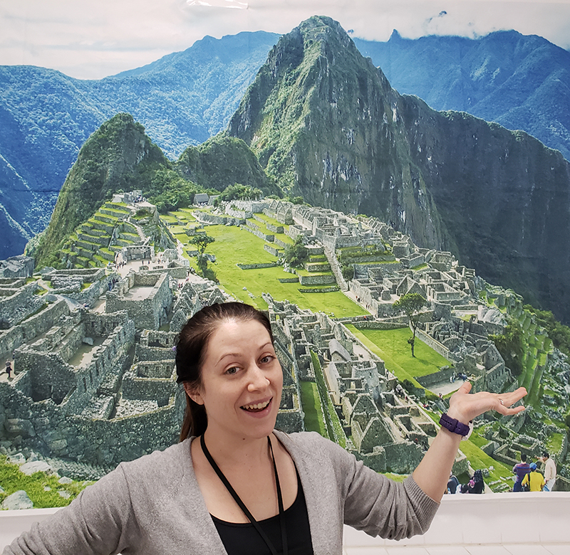 young woman with brown hair standing in front of a giant photo of the Incan ruins at Machu Picchu, Peru. There are tall, narrow mountains behind the stone ruins that are covered in green foliage.