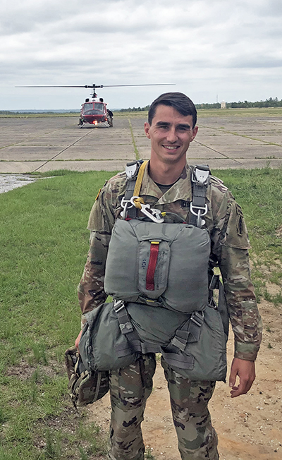 Young man with brown hair in camouflage army uniform with a parachute pack strapped on. There is a helicopter parked behdin him.