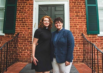 Photo of Gail and her wife Kathleen Davis standing on the steps in front of a red brick building with a green door and shutters. Gail is wearing her clerical collar and a black dress. Kathleen is wearing a blue patterned shirt, blue sportcoat, and khakis.