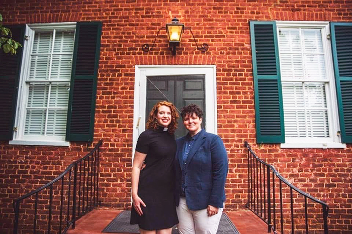 Photo of Gail and her wife Kathleen Davis standing on the steps in front of a red brick building with a green door and shutters. Gail is wearing her clerical collar and a black dress. Kathleen is wearing a blue patterned shirt, blue sportcoat, and khakis.