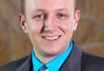 photo of Joe, smiling, wearing a gray coat, blue shirt, and plaid tie