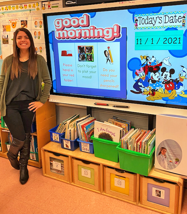 Photo of Kim with a big grin, standing beside a colorful classroom smartboard that says "good morning!" 