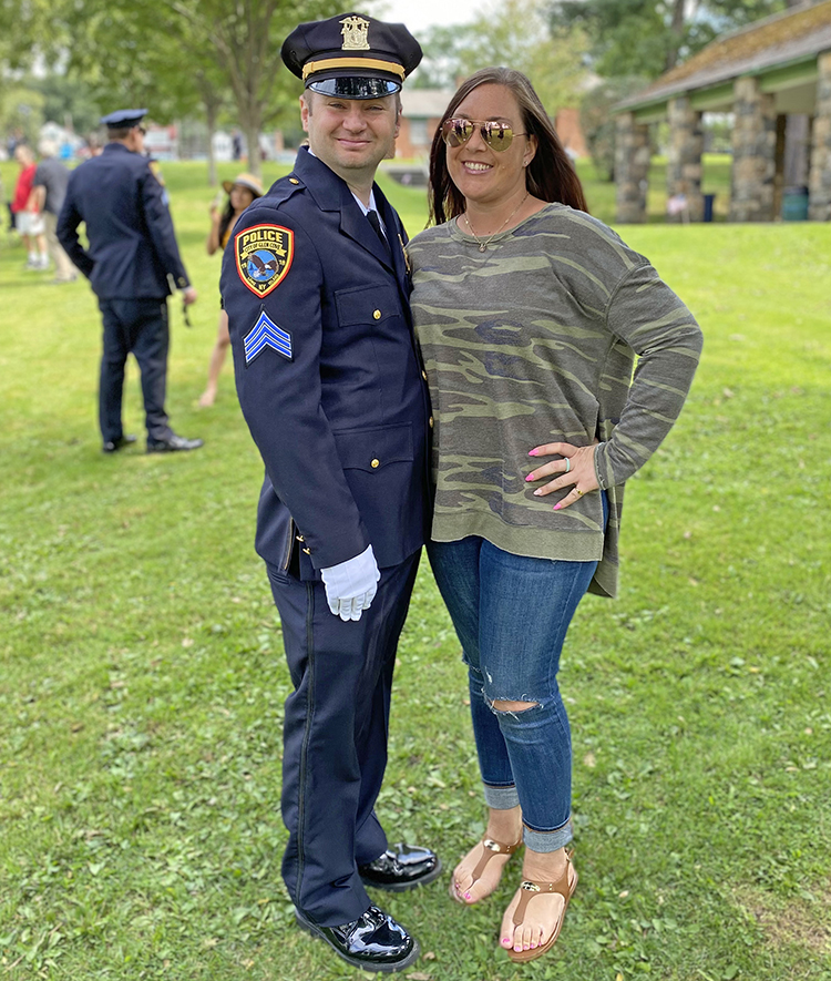 Photo of Matt standing in uniform, turned sideways so you can see the PD insignia and Sargeant stripes on his arm, he is posed with a woman in sunglasses, a long-sleeve t-shirt, and jeans who is also smiling 
