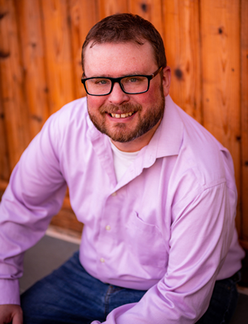 Photo of Ross, seating and smiling at the camera. He has red hair and a short beard, is wearing glasses and a purple button-down shirt.