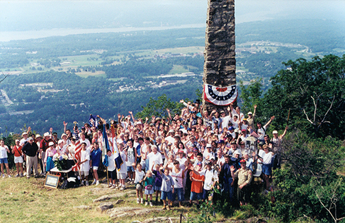 a crowd has gathered around a tall stone monument on the summit of Mount Beacon, you can see the city far below in the background