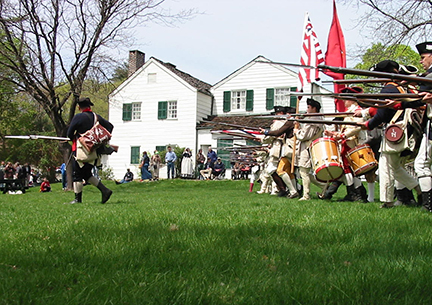 reenactors charge from the left to the right across a green lawn with bayonets fixed on their muskets, there is a white house in the background