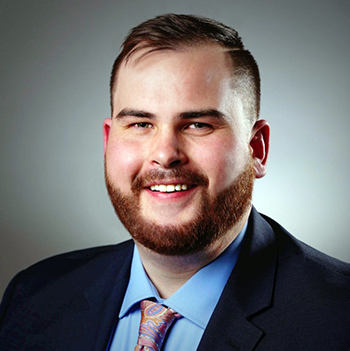 Photo of Brett, he has red hair and a beard, is wearing a dark blue coat, light blue shirt, and colorful paisley tie.