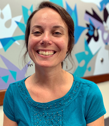 Photo of Colleen smiling. She has brown hair and is wearing a blue short. She stands in front of an abstract wall mural that is beige, blue, teal, black and white. 