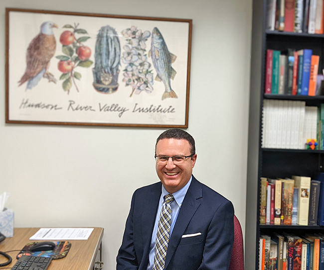 Photo of Dan sitting at a desk in his office. And HRVI poster hangs above his desk, and there is a book case full of history books beside him.