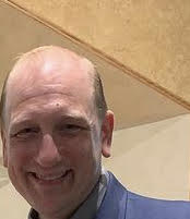 Snapshot of John, smiling. He has red hair and is wearing a blue coat over a gray shirt.