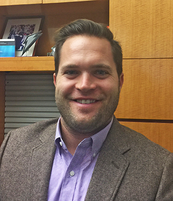 close up of John, smiling. He wears a purple shirt and a gray sportcoat and is in front of a wooden cabinet.