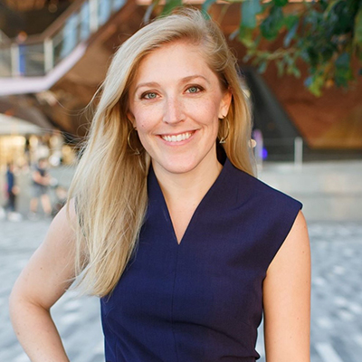 Photo of Lindsay, smiling, in a dark blue dress on a sunny day with a modern building in the blurred background.