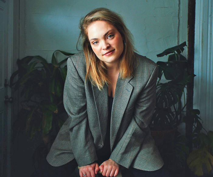 Photo of Marygrace, seated in a dimly lit blue room with plants behind her. She has blond hair, and is wearing a black and white plaid sportcoat.