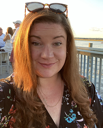 Photo of Mary Kate on a deck on the water. She is smiling, with her brows raised; it is a sunny day and the sky is blue.