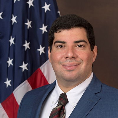 Close up photo of Michael, smiling in front of the American flag, he is wearing a light blue suit and a plaid tie.