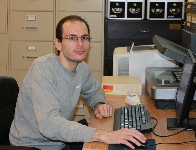Young man with short brown hair sitting at a desk in front of a computer. There are filing cabinets in the background.