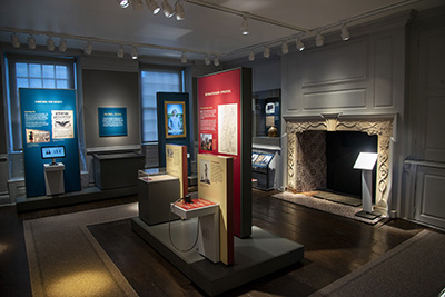 Historic, colonial, parlor with a carved stone fireplace, now set as a museum gallery with panels of text and images as well as artifacts.
