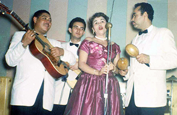 photo of a woman singing with one man playing guitar, one playing bass, and the third with maracas